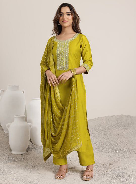 “Woman wearing a mustard Roman Silk embroidered kurta set with matching pants and dupatta, standing indoors.”