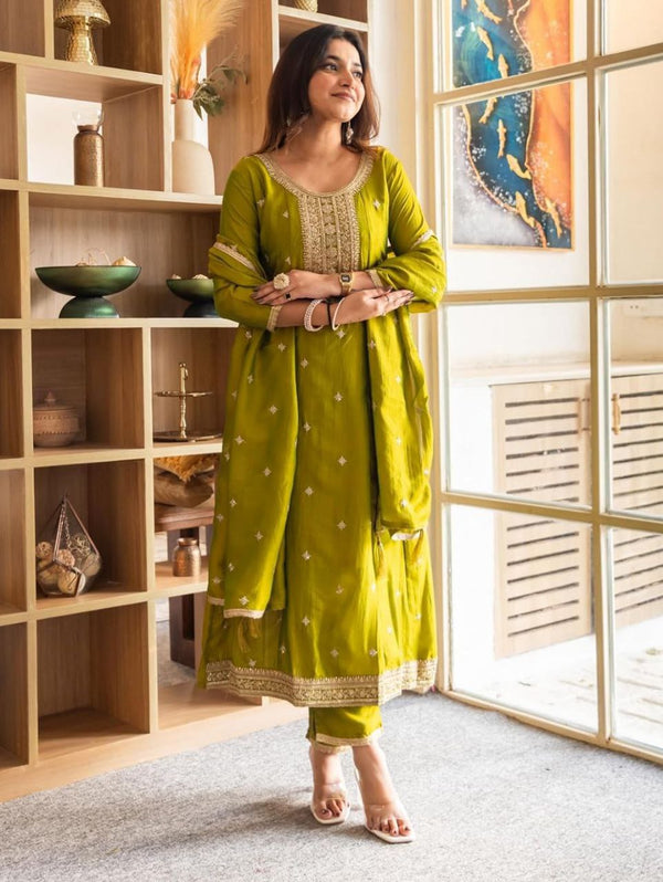 Woman wearing an olive-green embroidered Anarkali kurta set with matching pants and dupatta, standing indoors near a window.”