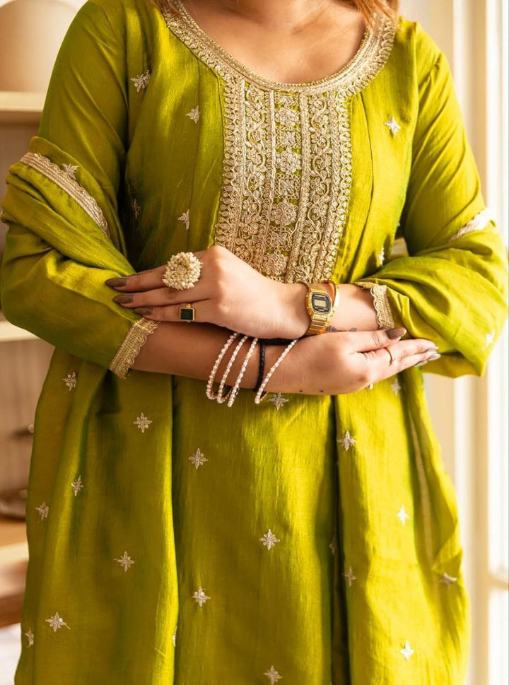 Side-angle lifestyle shot of a woman in an olive-green festive embroidered kurta set inside a modern living space.